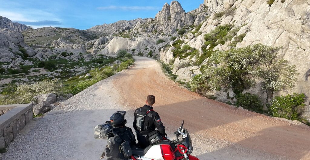 Essential Camera Gear for Motorcycle Travel Photography Fritz on Moto Guzzi V85 TT looking down a gravel road in Velebit Croatia — motorcycle travel photography Balkans